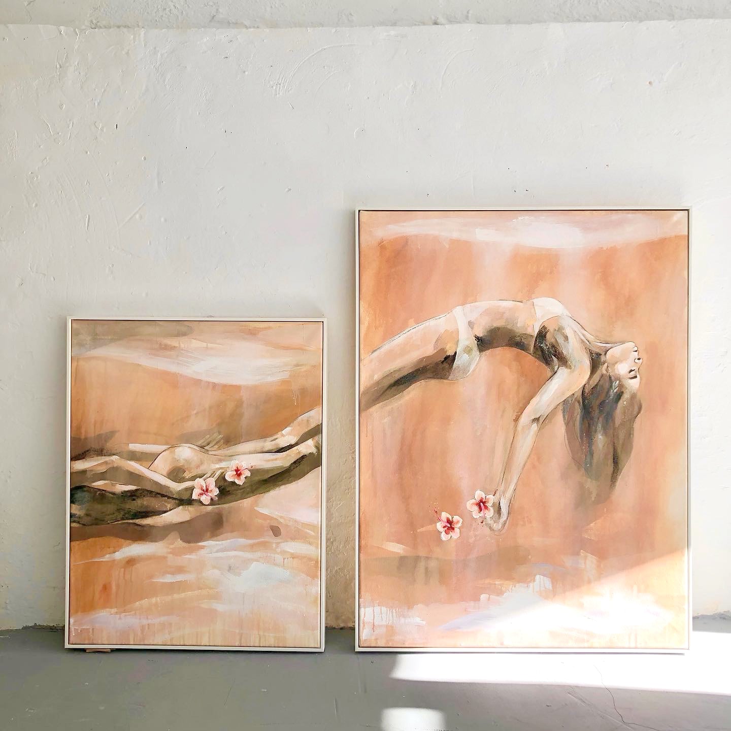 Two framed paintings leaning against a textured, light-colored wall. Both paintings share a warm, earthy color palette with soft brushstrokes and depict abstract female figures in fluid, serene poses. The larger painting on the right shows a woman in a deep backbend, her hair flowing downward while holding two delicate flowers. The smaller painting on the left portrays a reclining female figure, also holding flowers, blending into the soft background. The natural lighting and shadows add