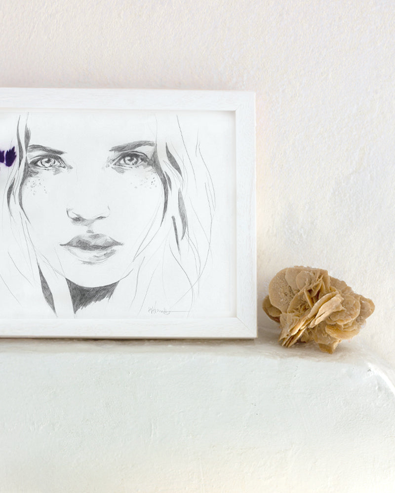 Woman pencil drawing in a white frame in an interior setting with a desert rose crystal.