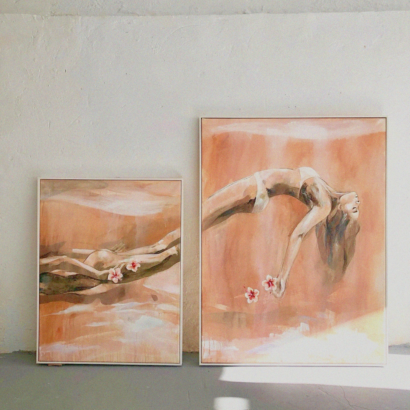 Two framed paintings leaning against a textured, light-colored wall. Both paintings share a warm, earthy color palette with soft brushstrokes and depict abstract female figures in fluid, serene poses. The larger painting on the right shows a woman in a deep backbend, her hair flowing downward while holding two delicate flowers. The smaller painting on the left portrays a reclining female figure, also holding flowers, blending into the soft background. The natural lighting and shadows add 