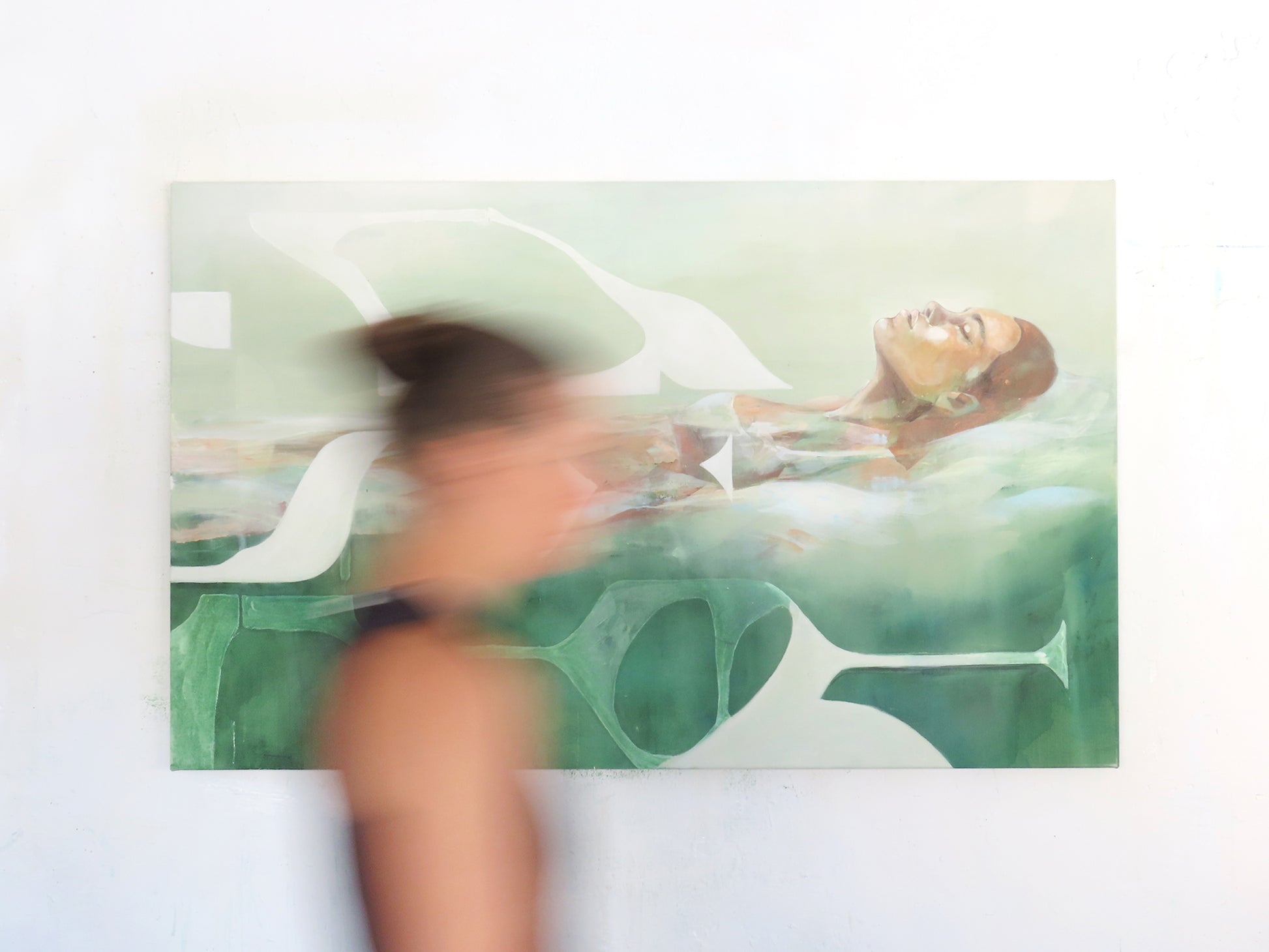 Photograph of a contemporary acrylic painting titled Mediterranean Musing by Hannah Adamaszek, featuring a serene woman floating in water, surrounded by abstract organic shapes in soft green and earthy tones. The painting is displayed on a white wall, with the Artist Hannah Adamaszek walking past.