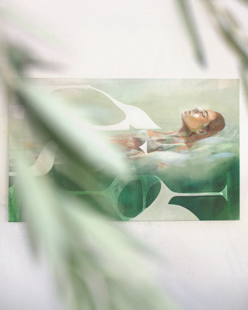 Photograph of a contemporary acrylic painting titled Mediterranean Musing by Hannah Adamaszek, featuring a serene woman floating in water, surrounded by abstract organic shapes in soft green and earthy tones. The painting is displayed on a white wall, partially obscured by blurred green foliage in the foreground, adding depth and a natural ambiance.