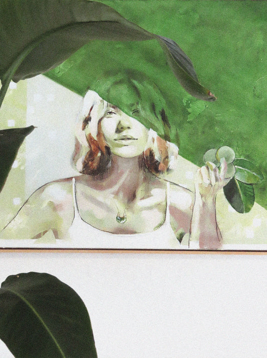 Painting Nourish by Hannah adamaszek of a bohemian woman in green at one with nature. Leaves are at the front of the photo with the framed artwork behind.